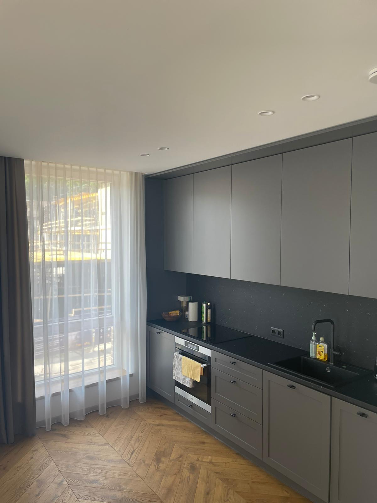 Virtuvė su pilkomis spintelėmis, juodais stalviršiais ir dideliu langu.