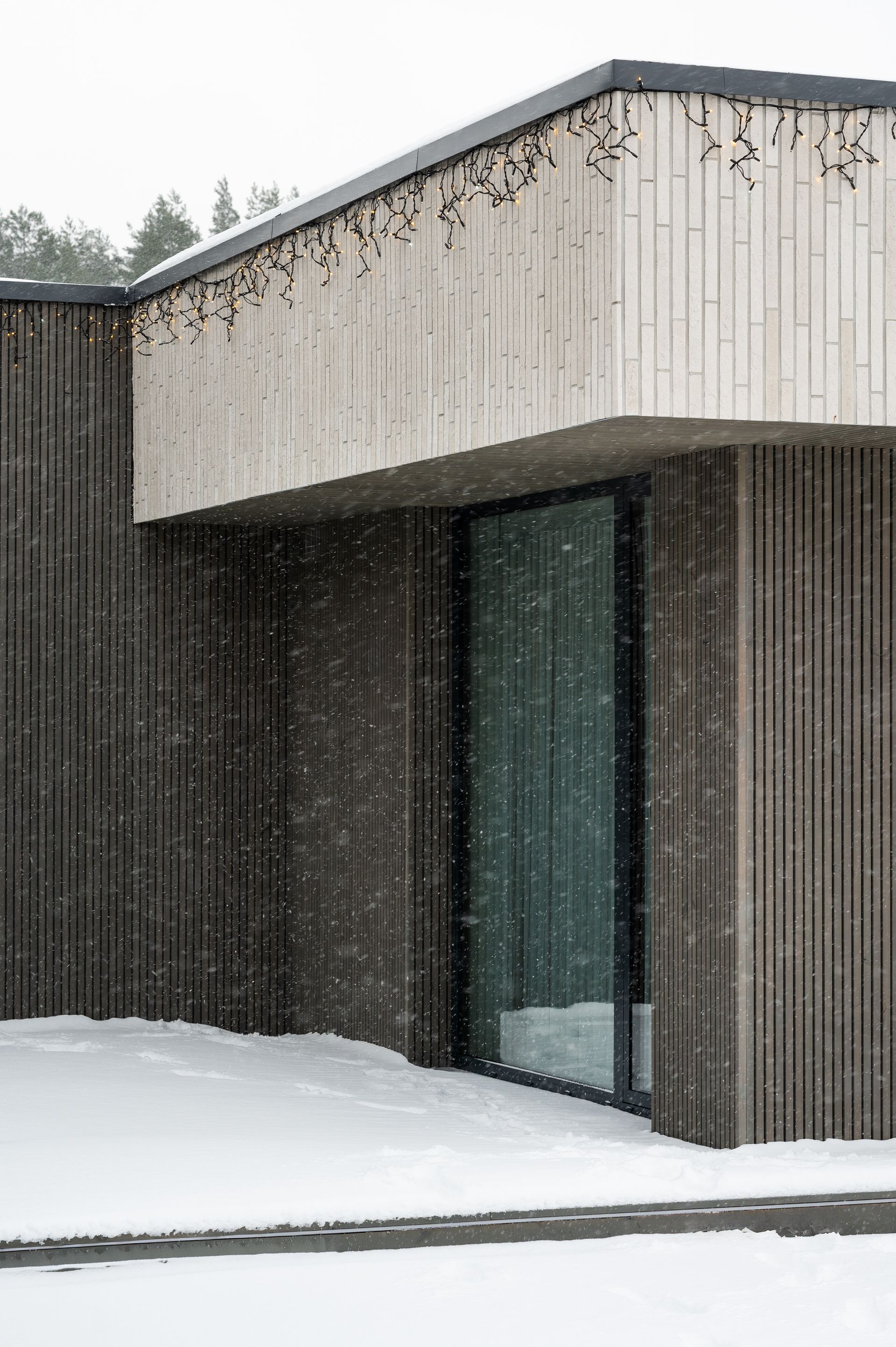 Modern building with dark wood siding, concrete accents, and a large glass window. Snow falls on the snowy ground.