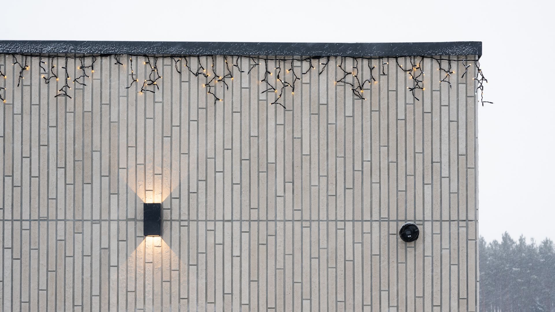Exterior wall of a building with vertical brick-like texture. It has a wall-mounted light and a round black vent. Icicle lights are draped from the roof.