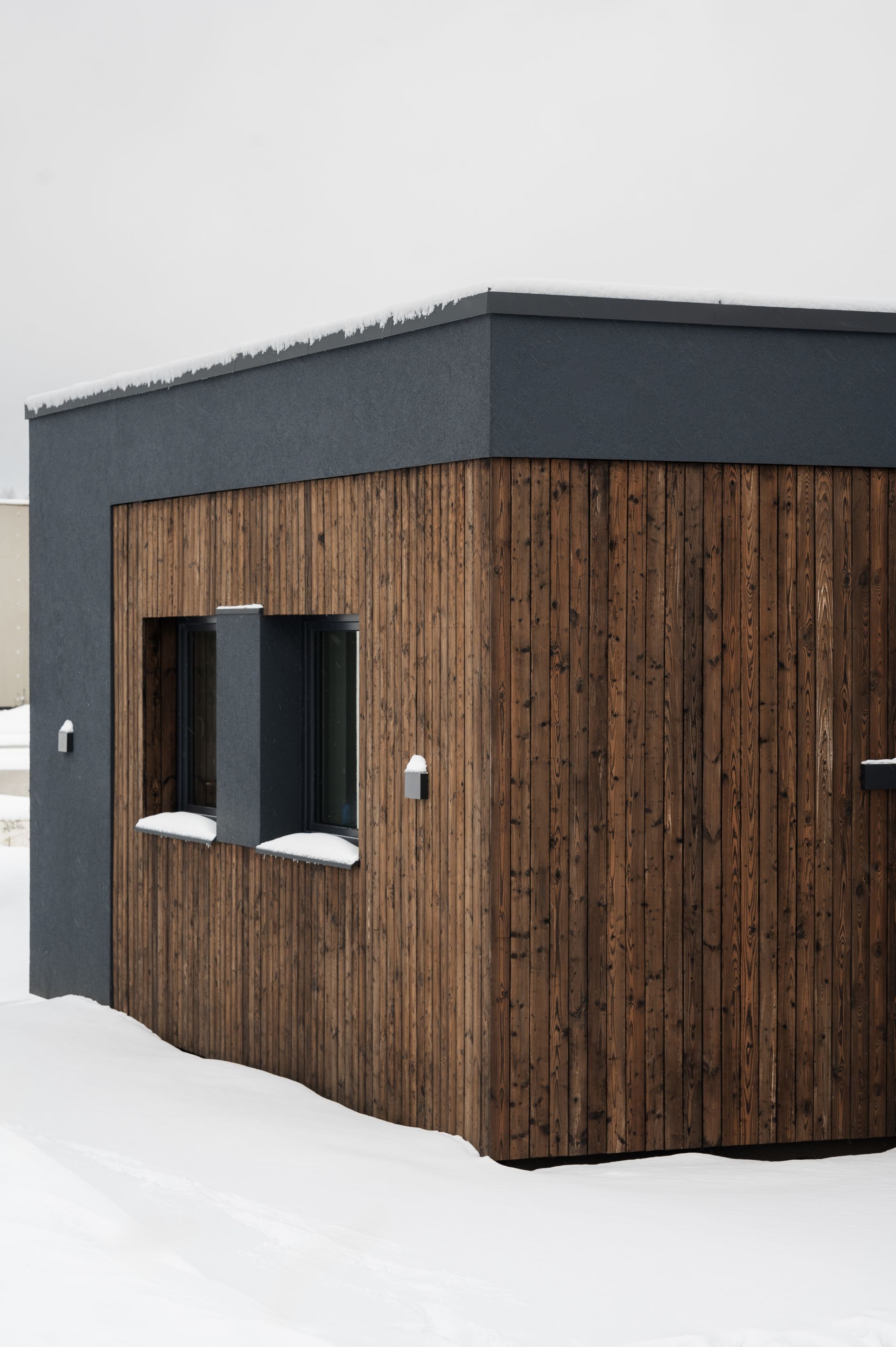 Modern wooden building with dark trim, two windows, and snow-covered ground.
