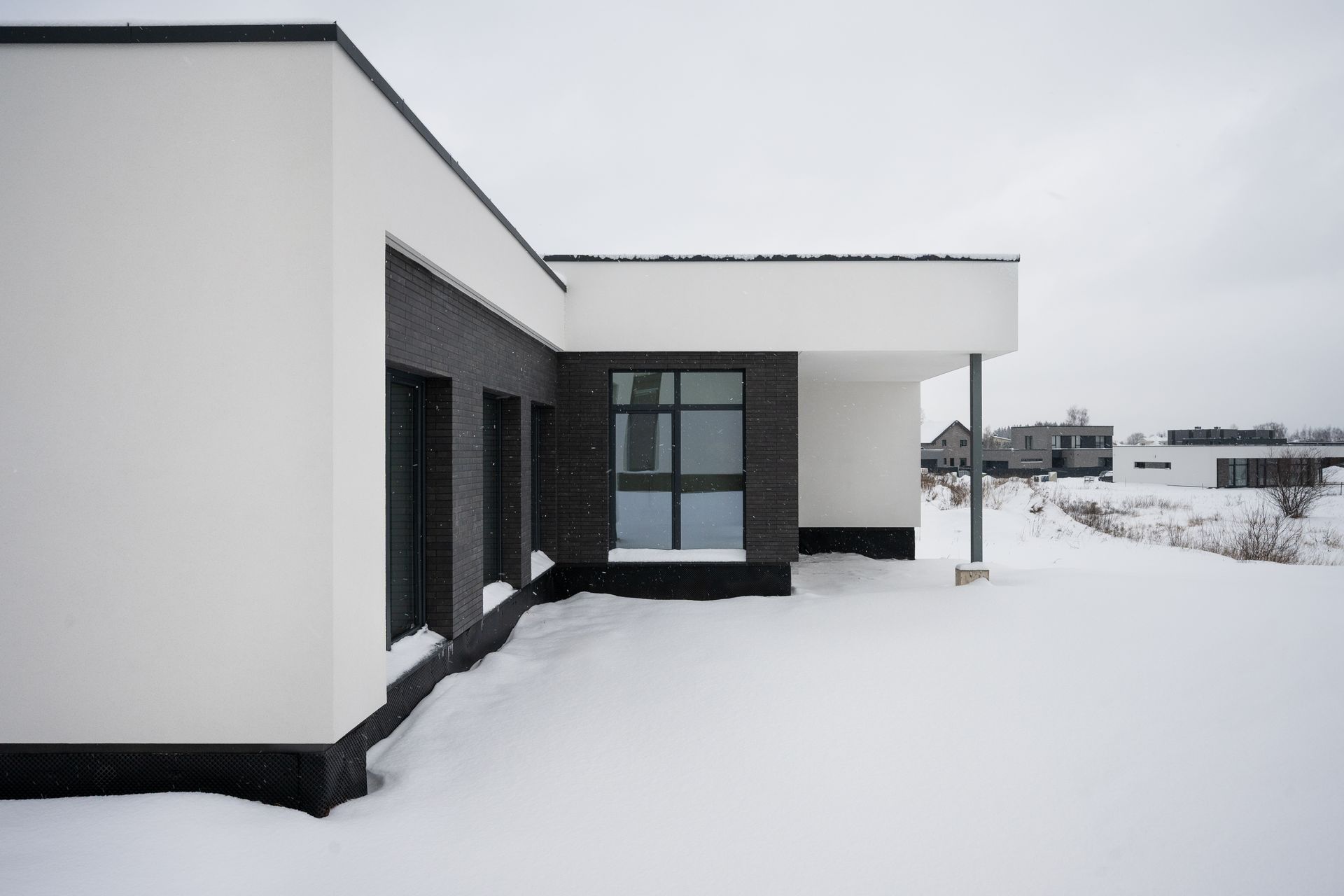 Modern white and black house exterior with snow-covered ground, overcast sky.