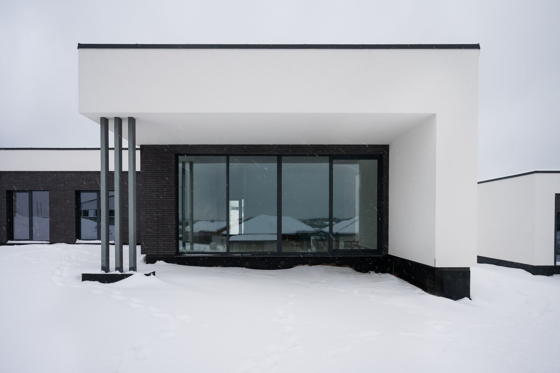 Modern house exterior with large windows, black brick, and white accents, in a snowy setting.