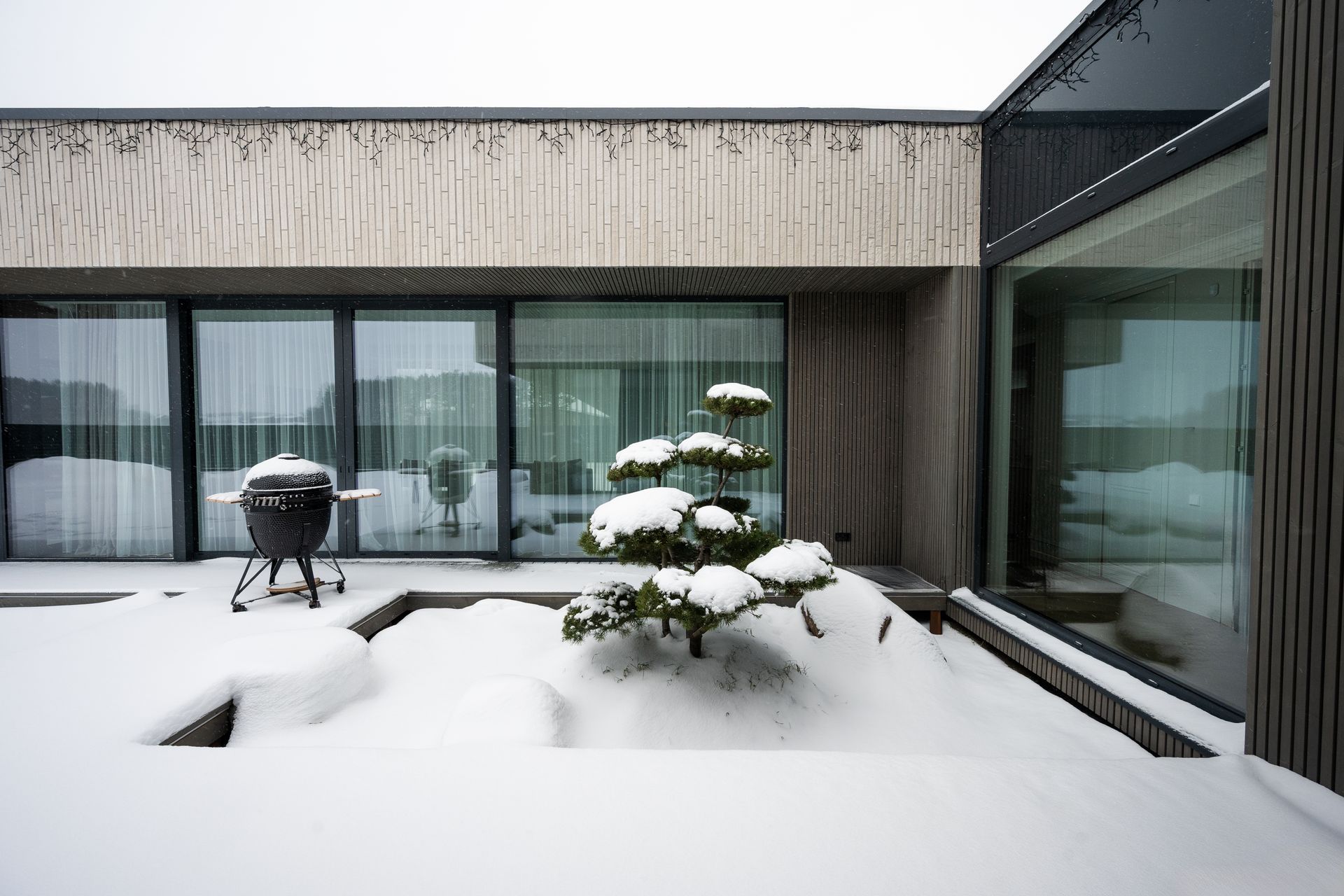 Snow-covered outdoor patio with a small tree and grill in front of a modern building with large windows.