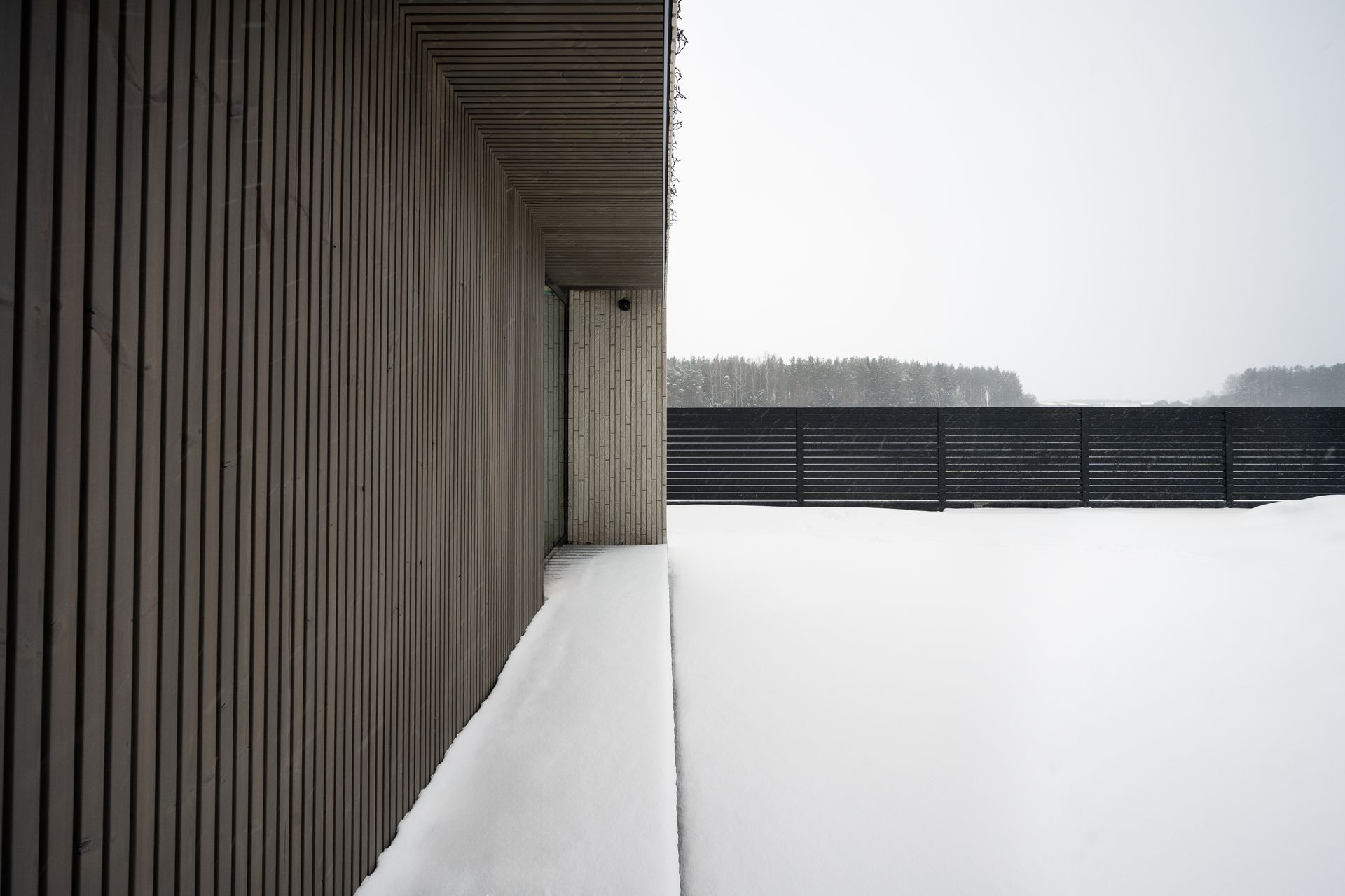 Snow-covered balcony, wooden wall on the left, snow-filled landscape on the right, gray sky.