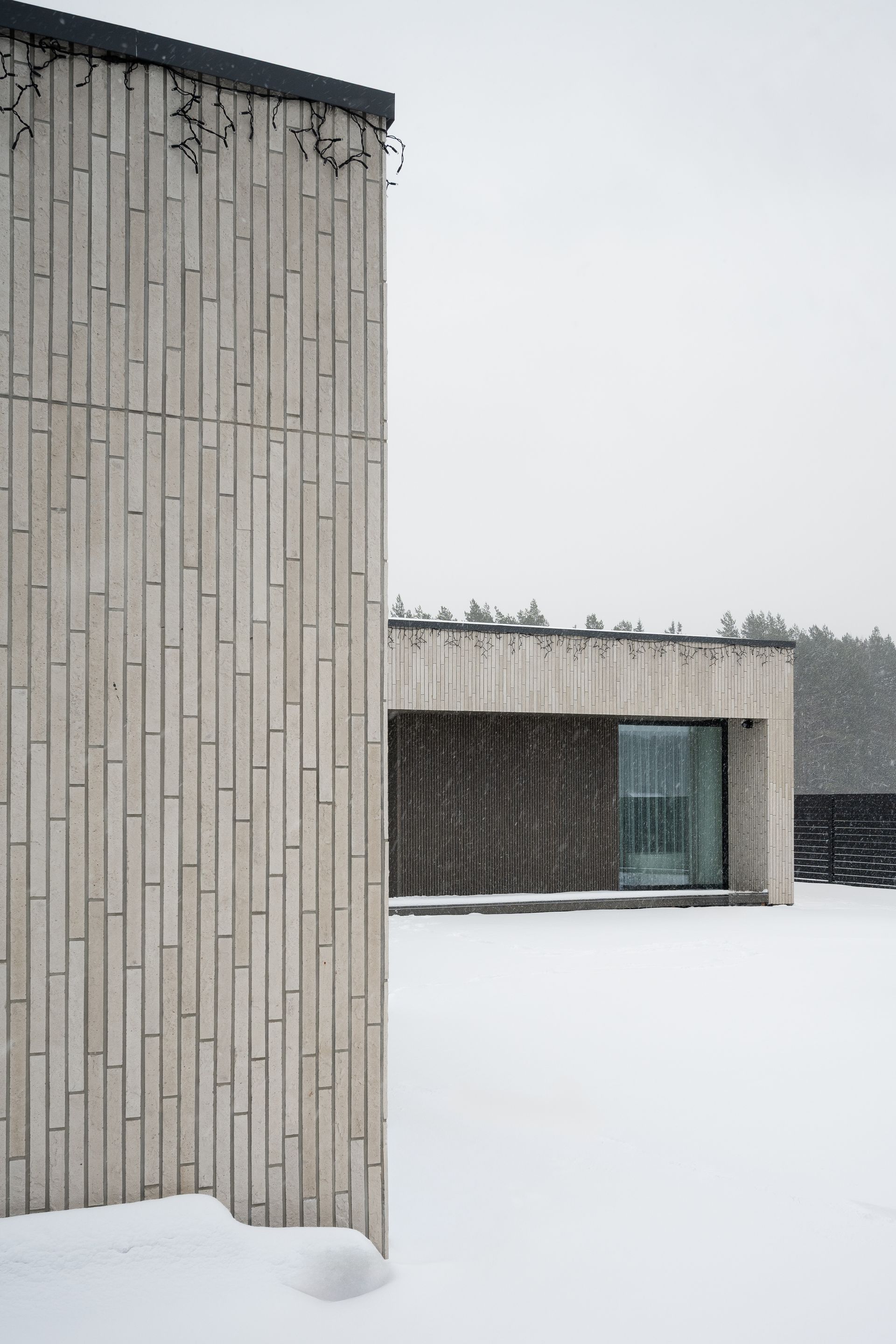 Modern, light-colored wooden buildings in snowy landscape. One building is in the foreground, another is further back. Snow falling.