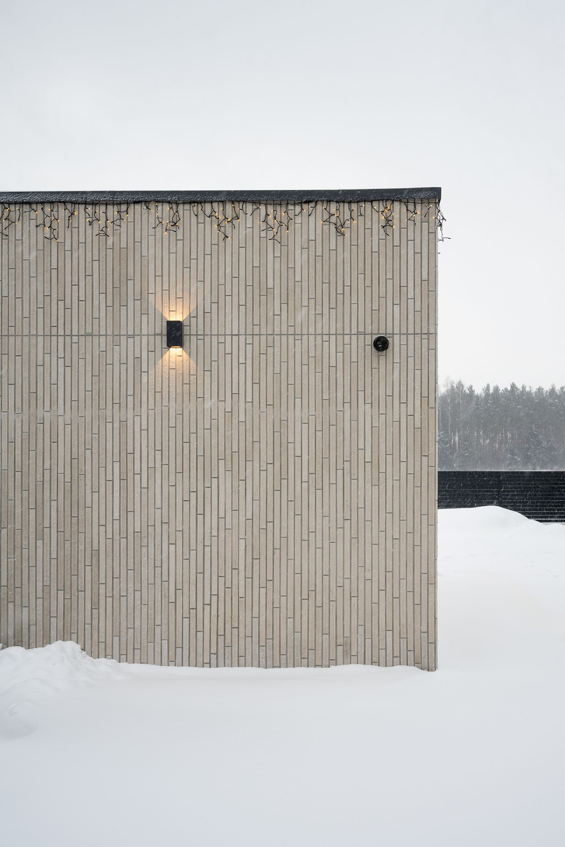 Snowy exterior of a building with light-colored, vertically striped facade, two wall lights, and a dark roof edge.