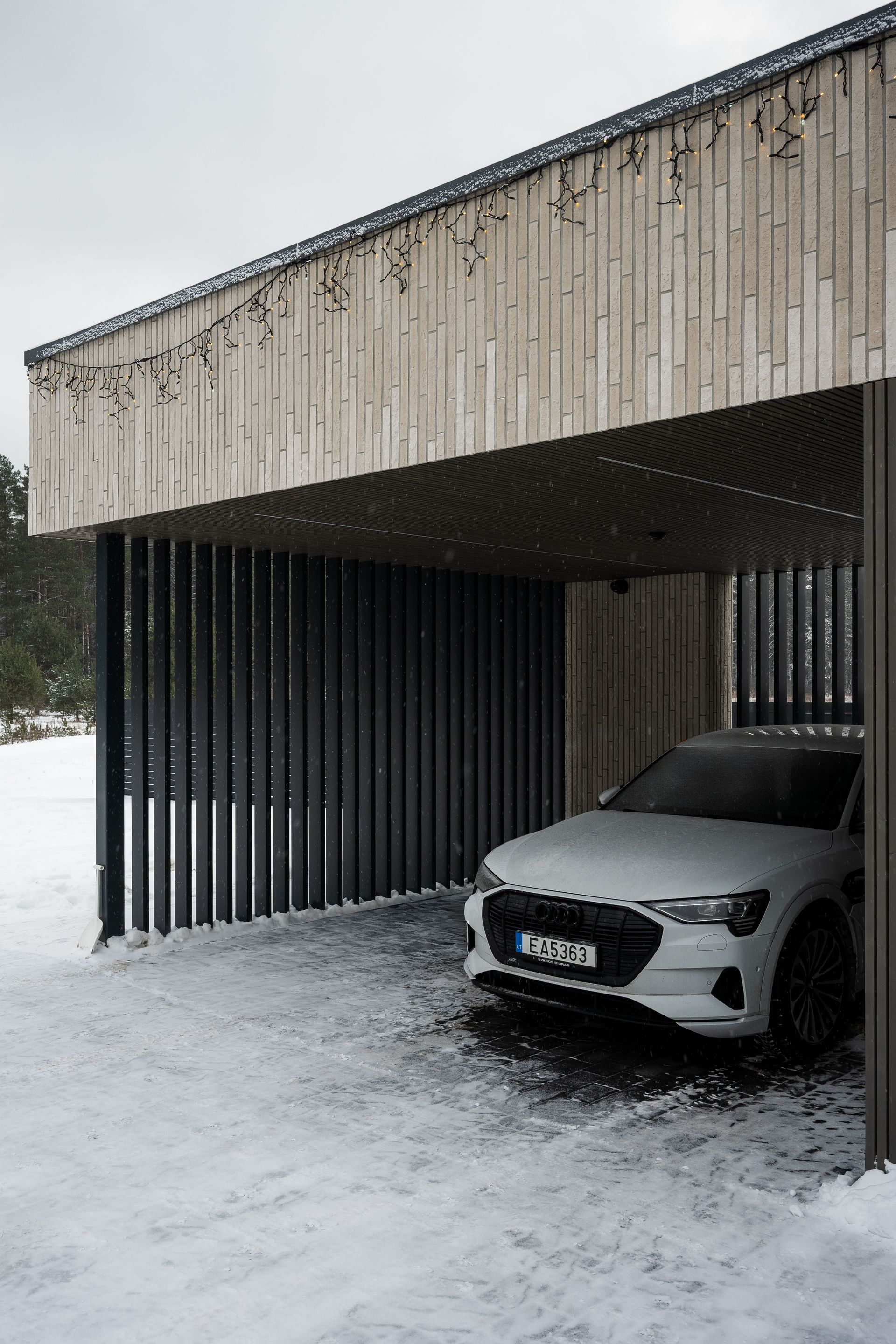White car parked under a carport with vertical slats, snow falling.
