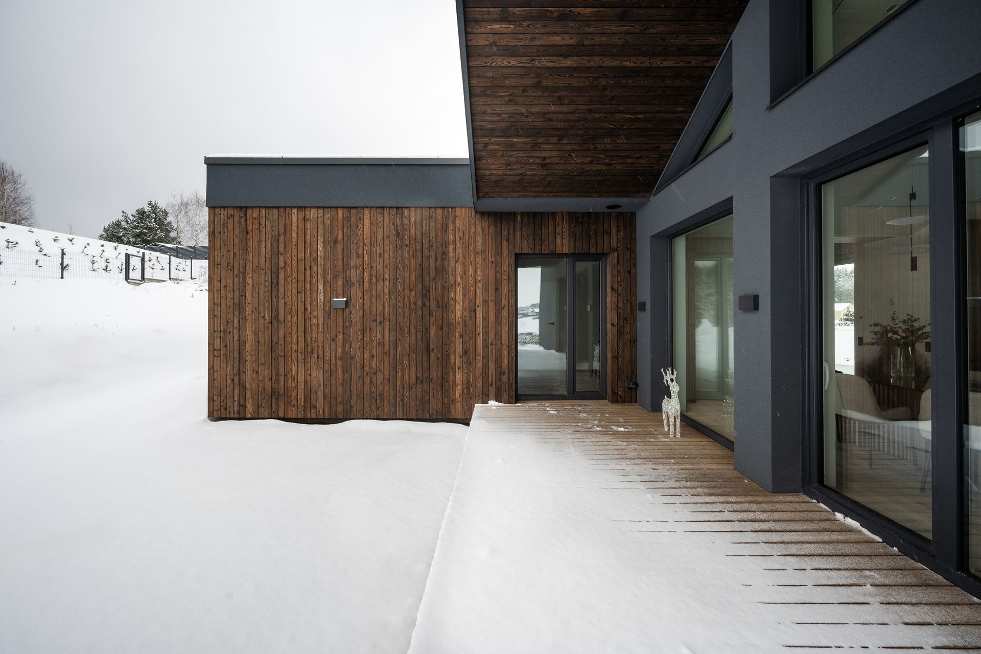 Snow-covered exterior of modern house. Wood siding, grey walls, and large windows.