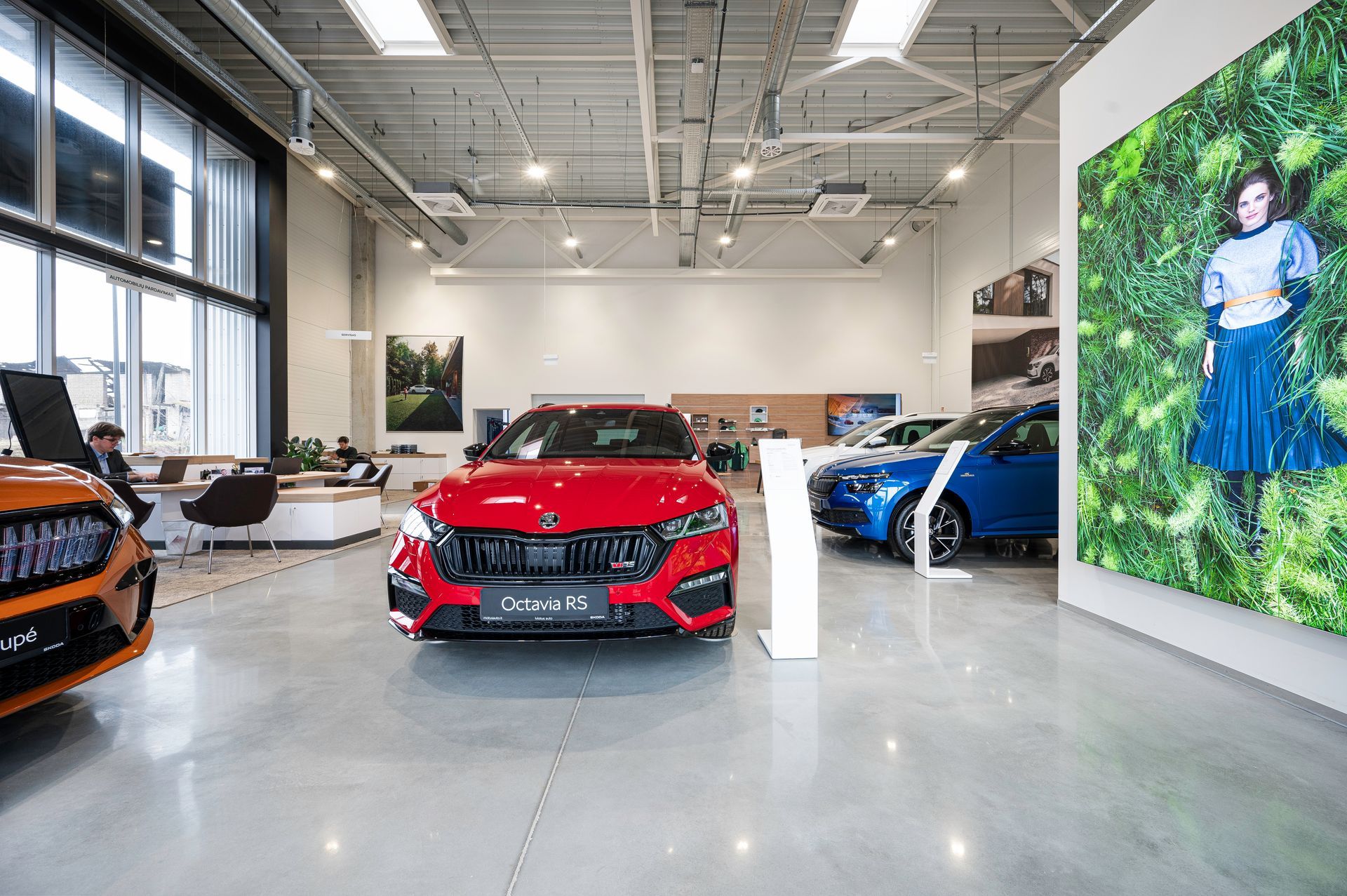 Raudonas automobilis stovi salone šalia mėlyno automobilio.