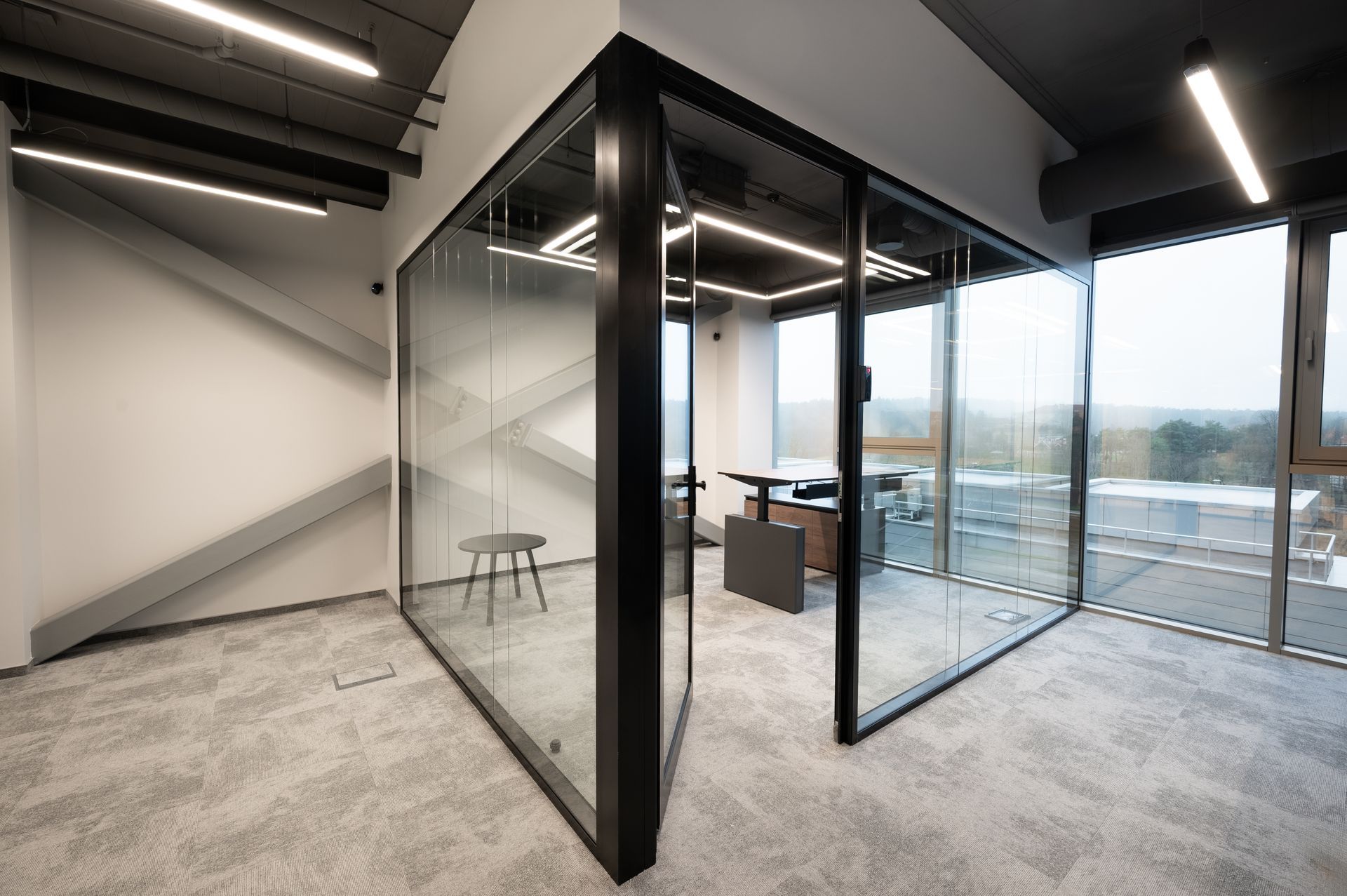 Modern office with glass-walled office space, a desk, and a small chair. Gray carpet and exterior view through windows.