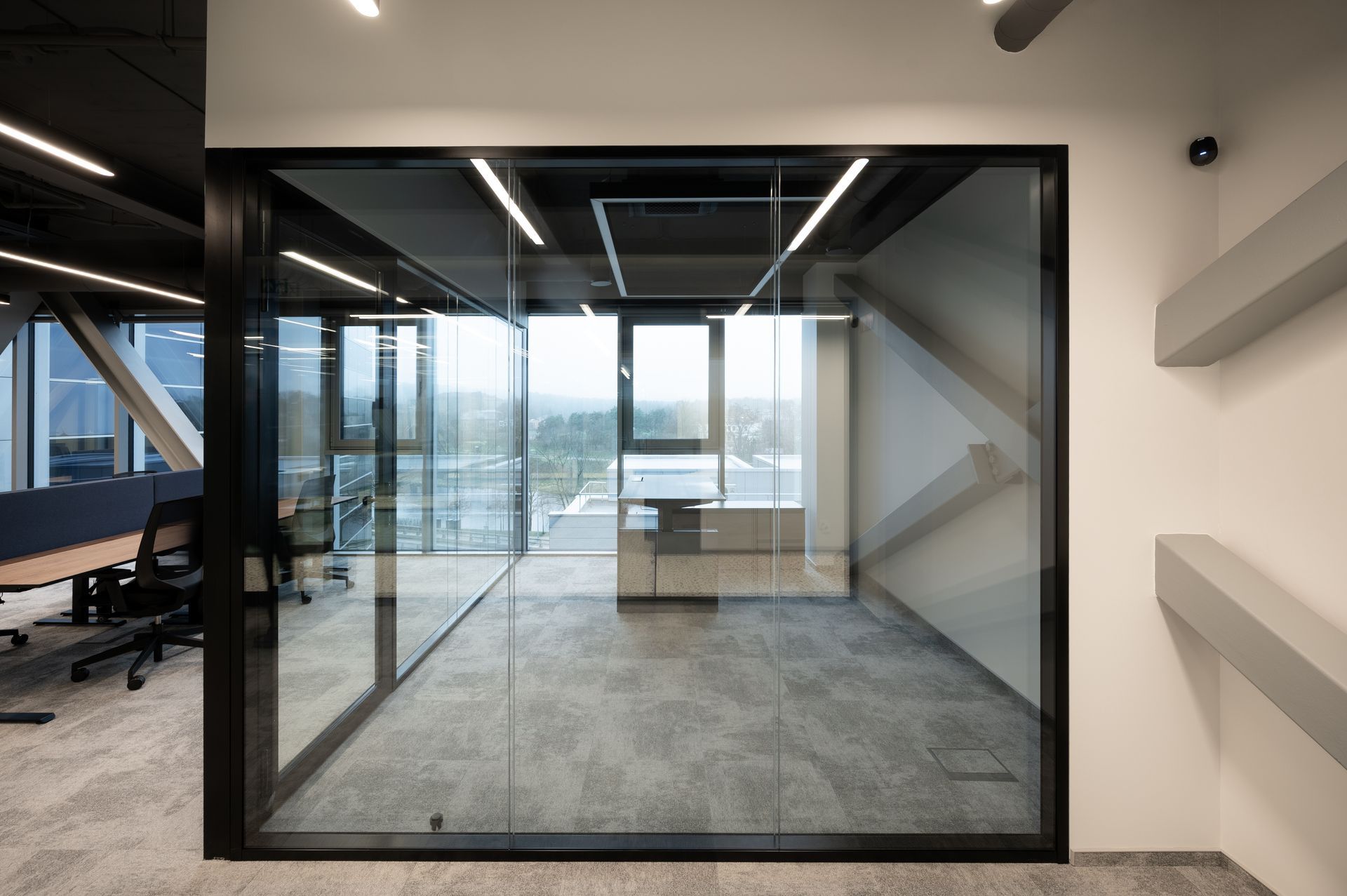 Glass-walled office with black trim. Gray carpet, white walls. View through window of outside landscape.