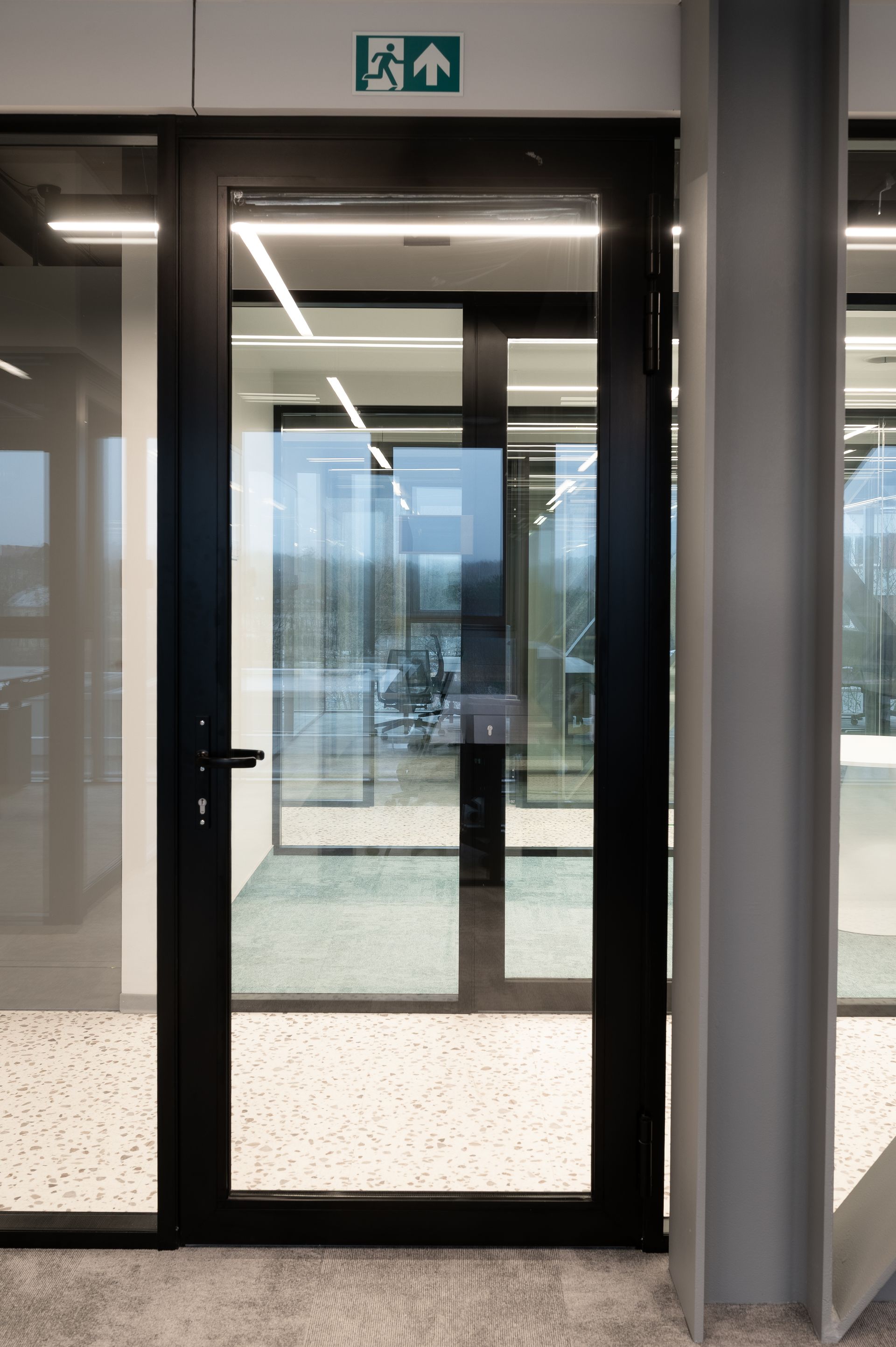 Black-framed glass door with a green exit sign above. Located in a modern interior, seen on a speckled floor.