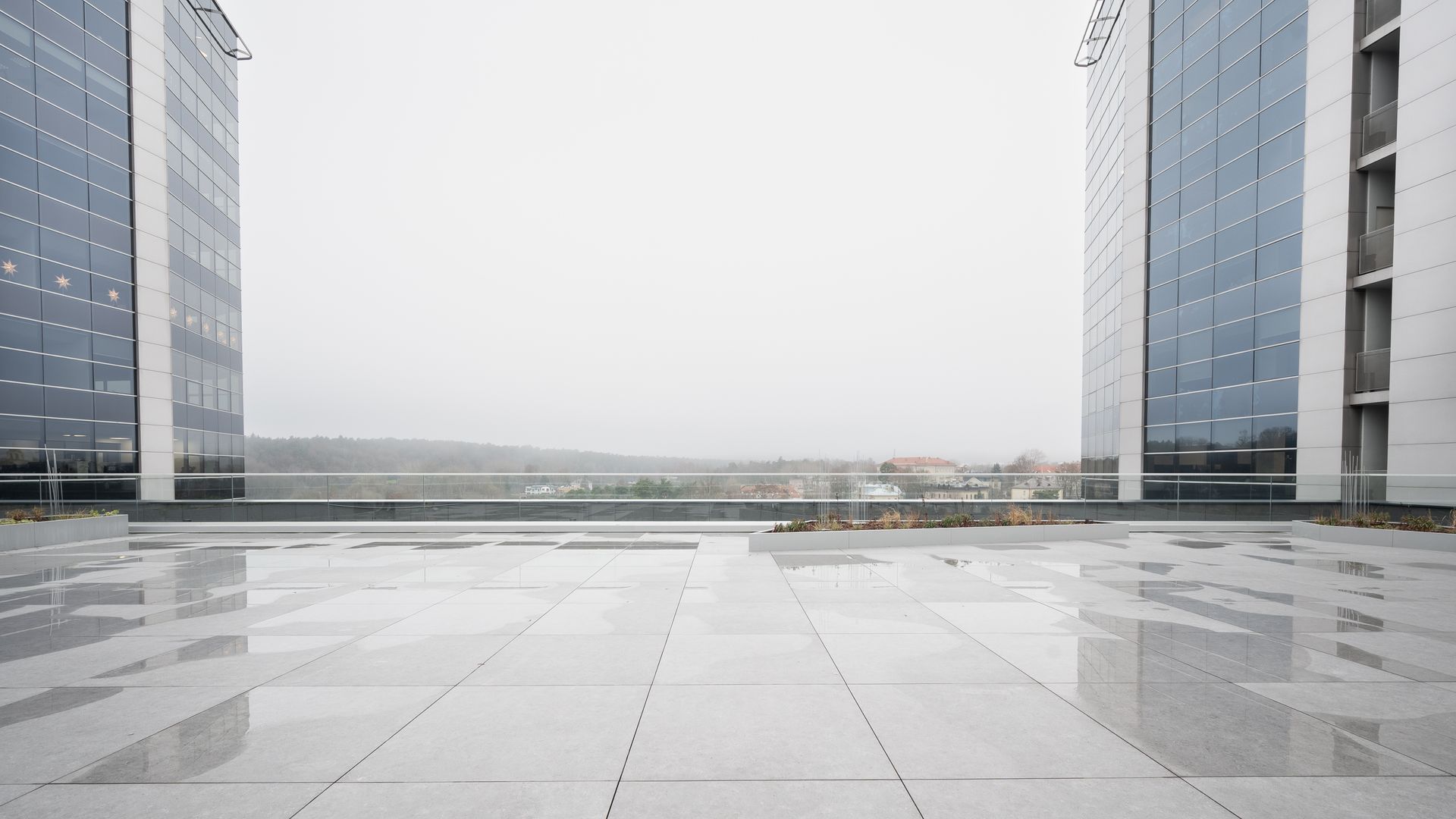 Two modern buildings flank a wet plaza on a cloudy day, distant landscape visible.
