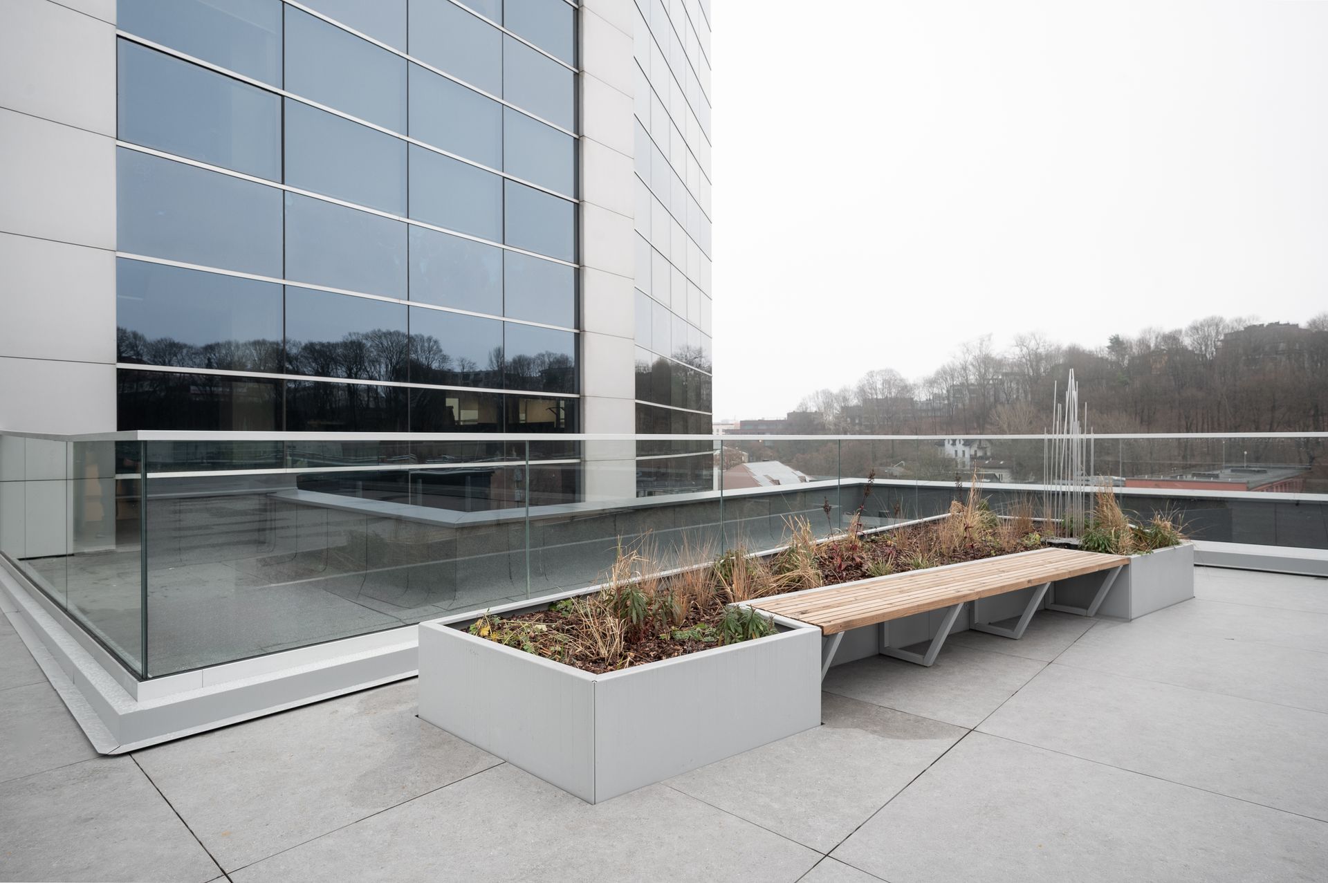 Modern building rooftop with a bench and planters. Gray, glass building exterior in background. Overcast day.