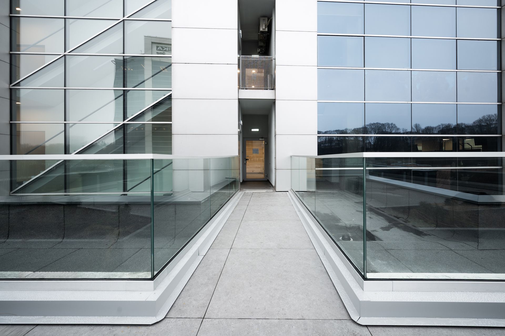 Hallway with glass walls and metal railings, leading to an open doorway. Gray and white.