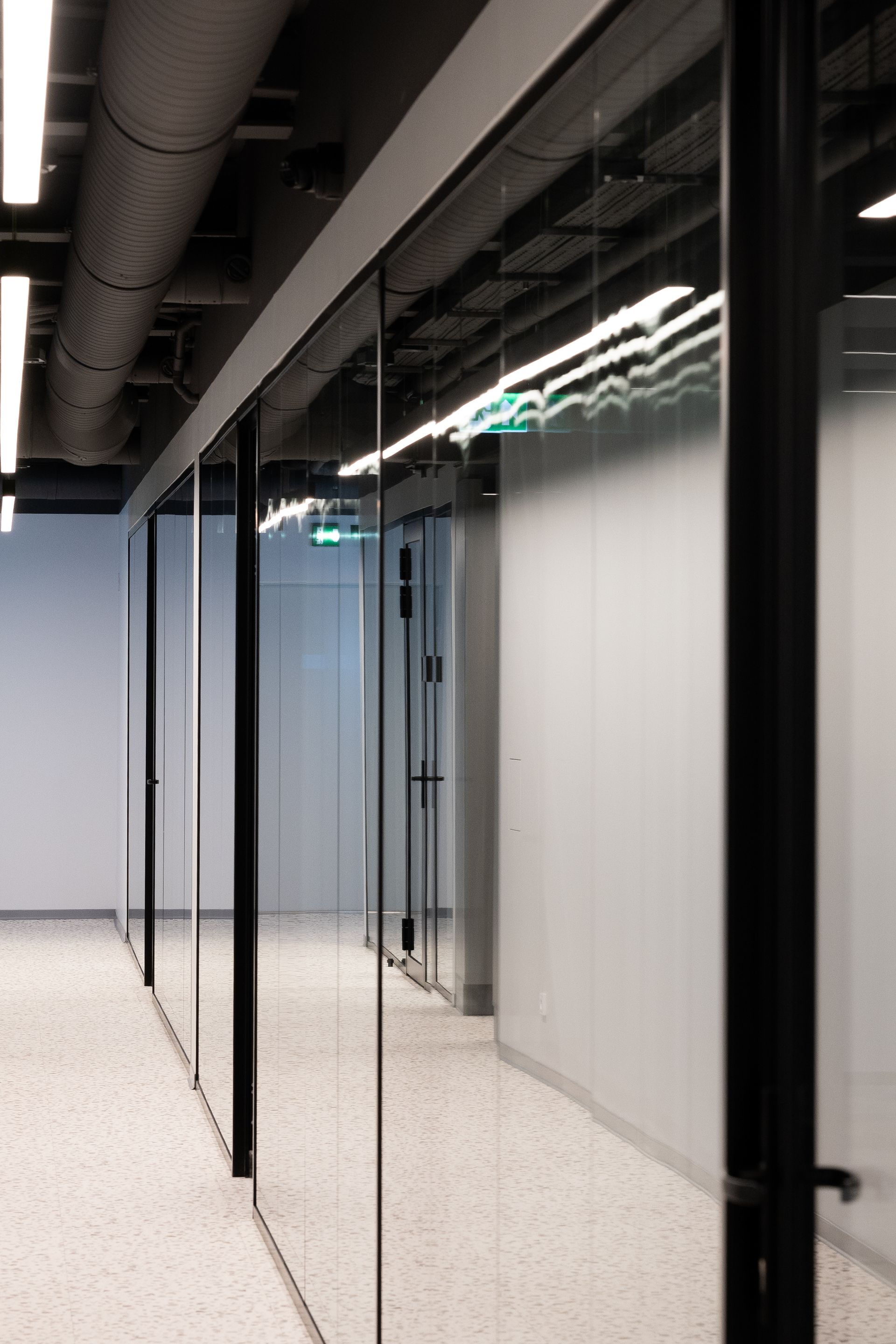 Modern office hallway with glass walls and black frames, reflecting ceiling lights.