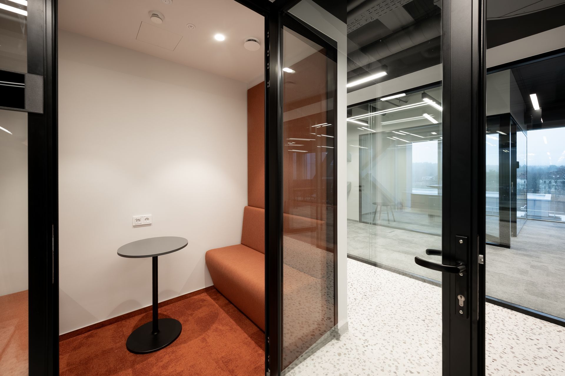 Office phone booth with orange seating, small table, glass walls, and black frame.