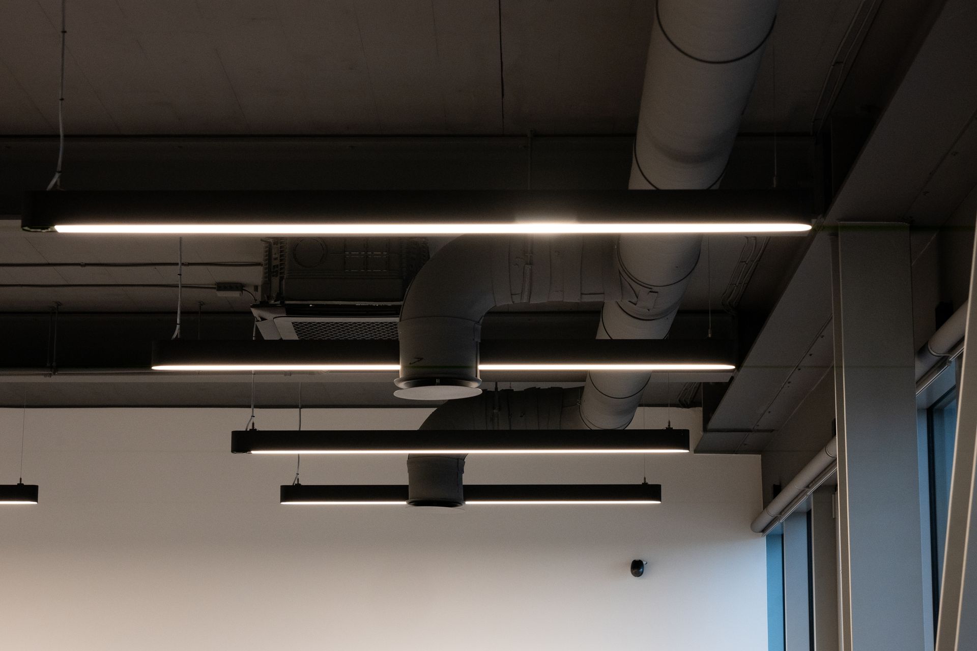 Overhead view of a modern office space with linear lights suspended from a concrete ceiling. Ventilation ducts are also visible.