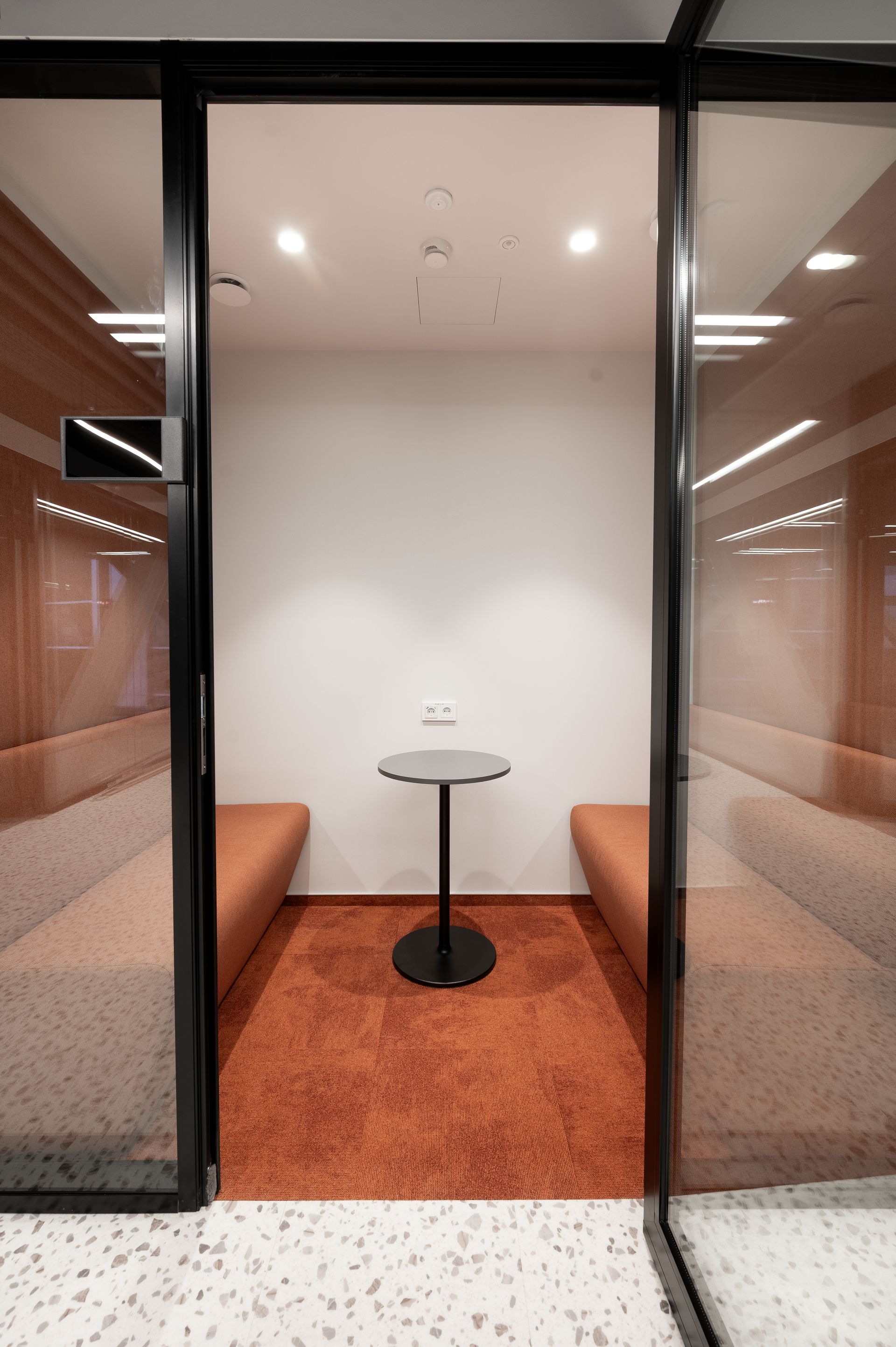 Acoustic booth with orange carpet and seating, small table, and glass walls with black frame.