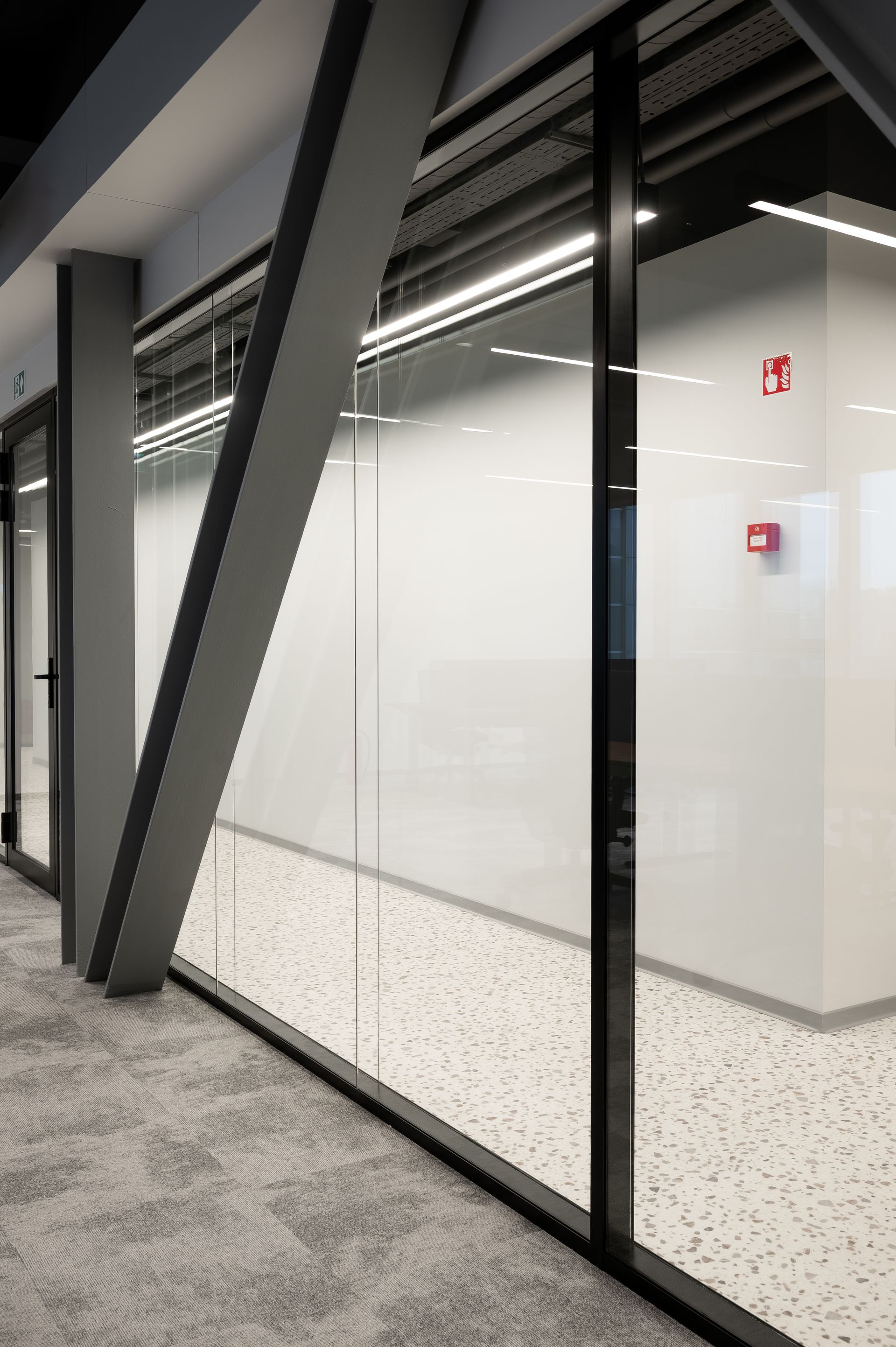 Interior office hallway with glass walls, gray support beam, and patterned carpet.