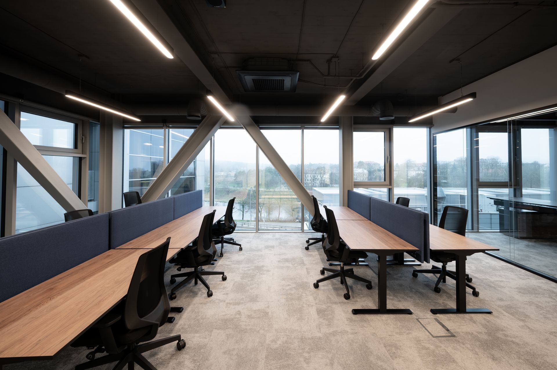Modern office workspace with desks, chairs, and large windows. Black ceiling, neutral carpet, and angled support beams.
