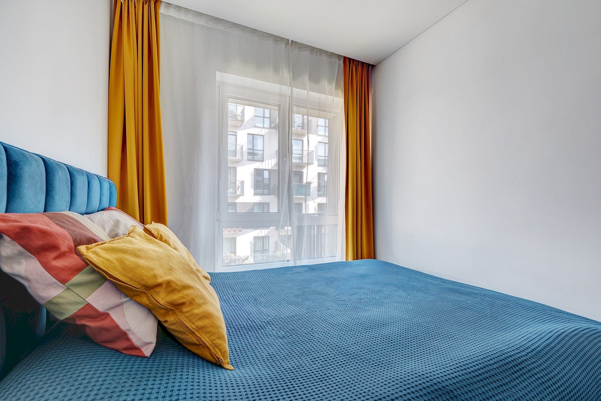 Miegamasis su mėlyna lova, geltonomis užuolaidomis ir langu.