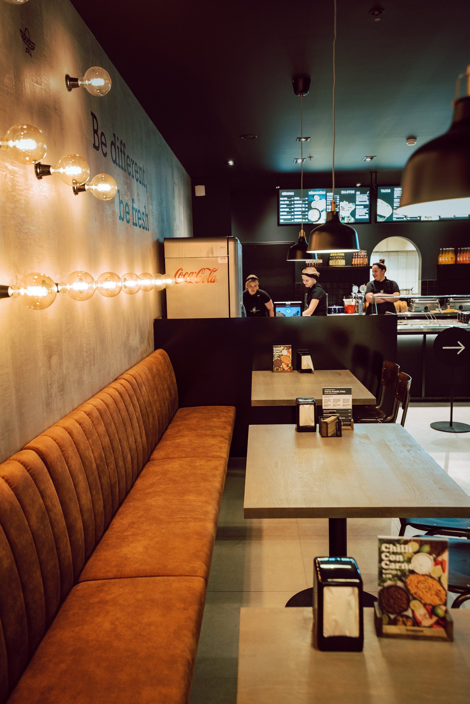 Restoranas su stalais, suolais ir fone stovinčiais žmonėmis.