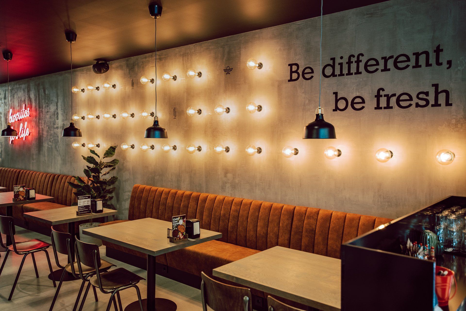 Restoranas su stalais, kėdėmis ir užrašu ant sienos „būk kitoks, būk šviežias“.