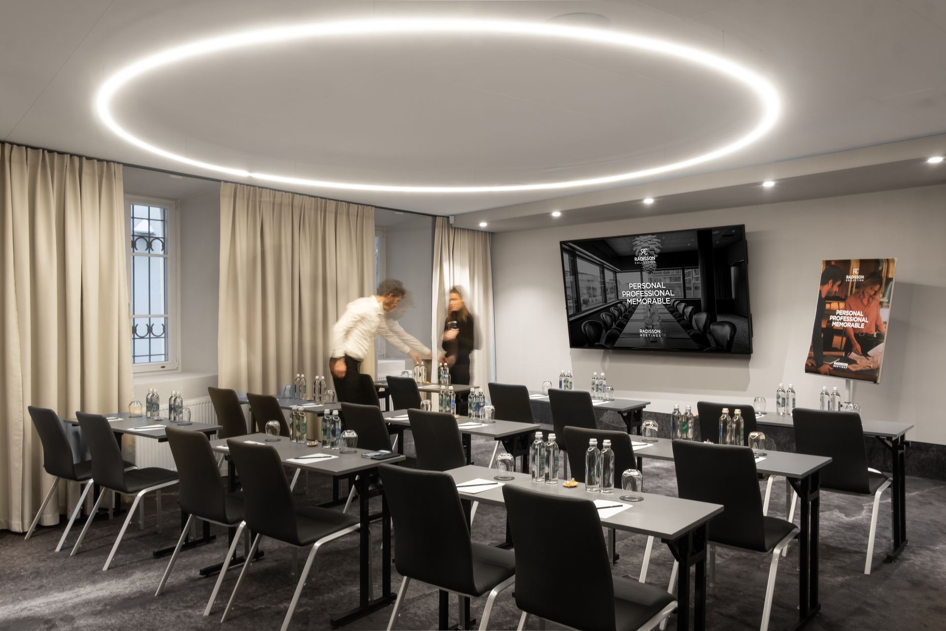 Konferencijų salė su stalais, kėdėmis ir vyras, stovintis tarpduryje.