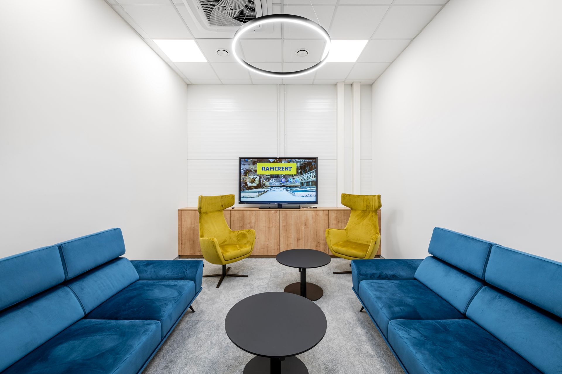 Svetainė su mėlynomis sofomis, geltonais foteliais ir plokščiaekraniu televizoriumi.