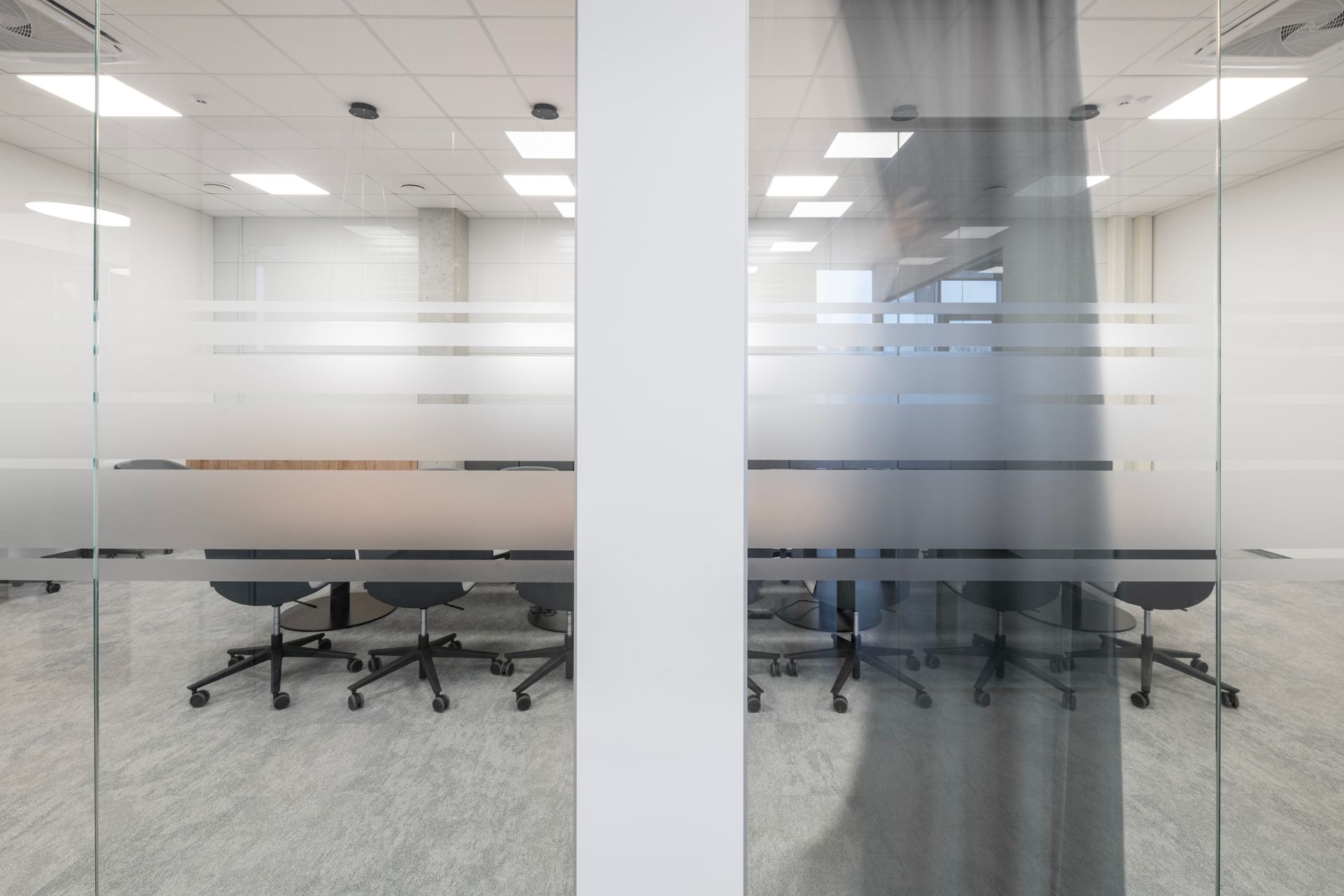 Konferencijų salė su ilgu stalu ir kėdėmis už stiklinių durų.