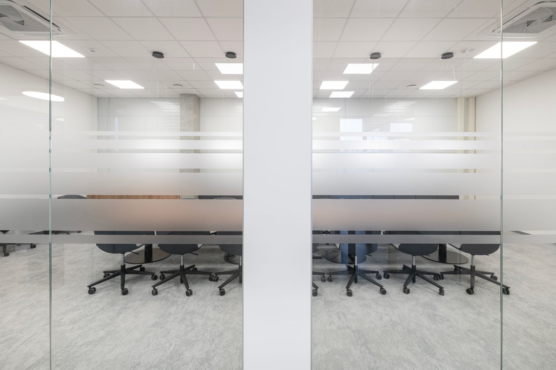 Konferencijų salė su ilgu stalu ir kėdėmis už stiklinių durų.