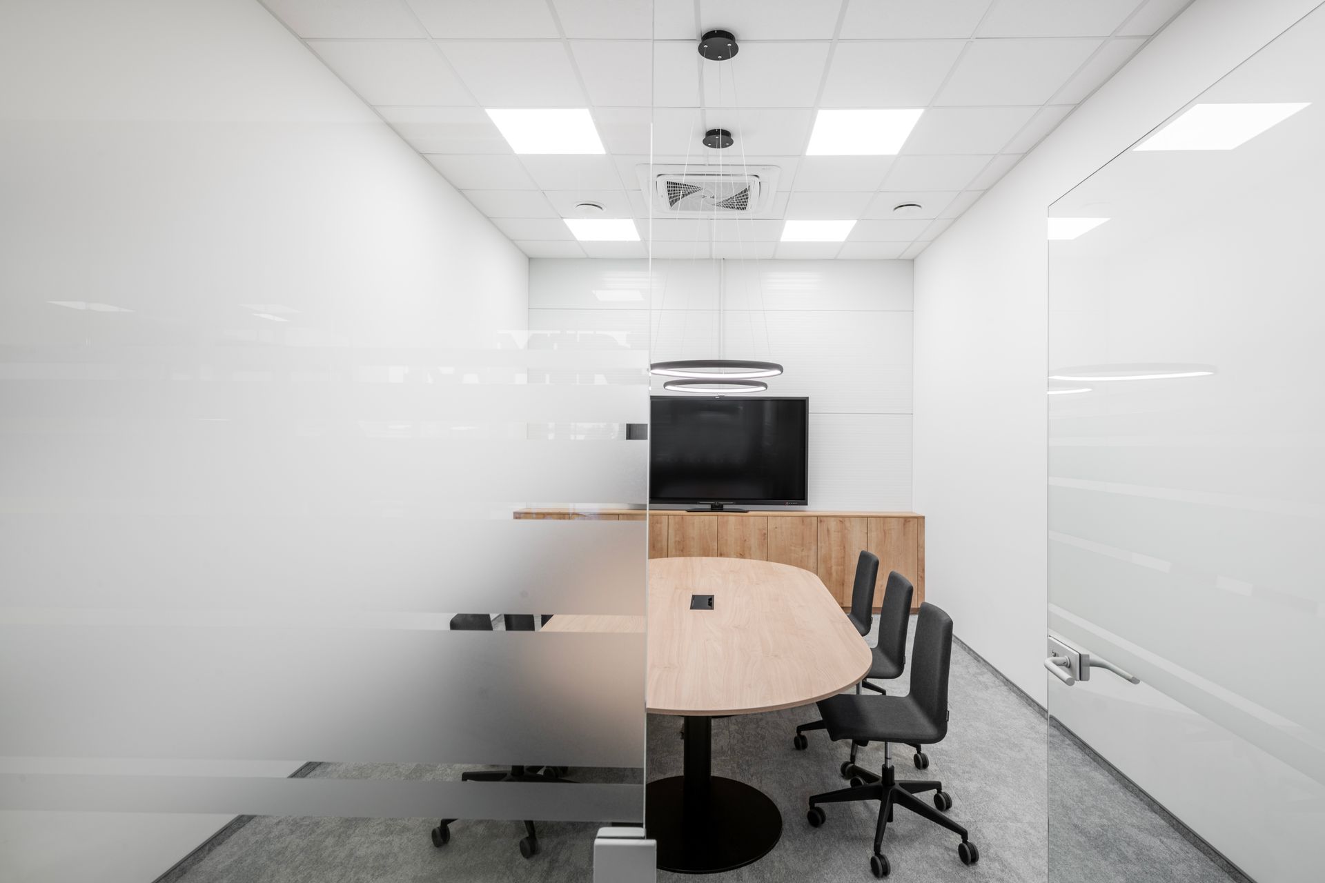 Konferencijų salė su stalu, kėdėmis ir televizoriumi.