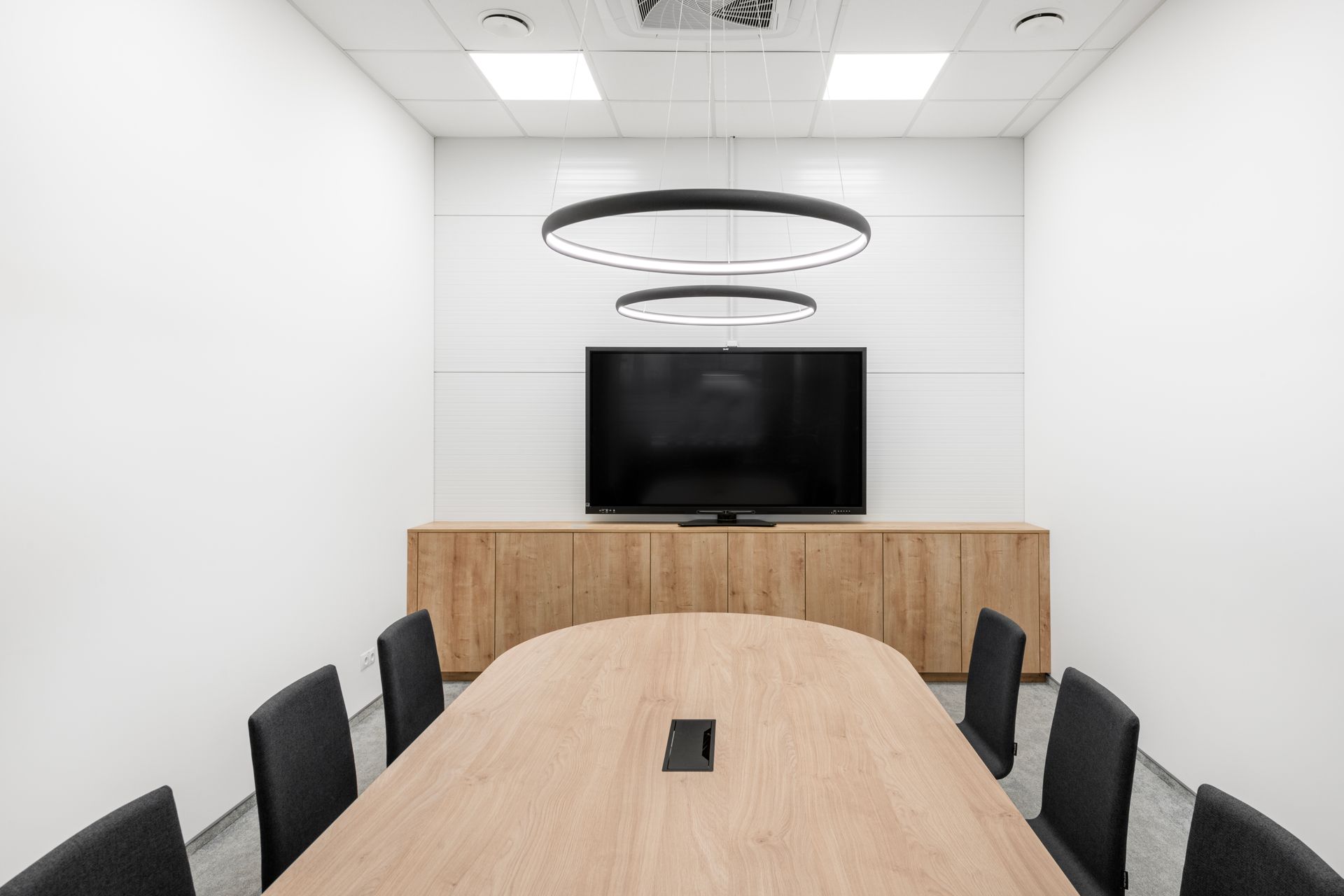 Konferencijų salė su ilgu mediniu stalu, kėdėmis ir plokščiaekraniu televizoriumi.