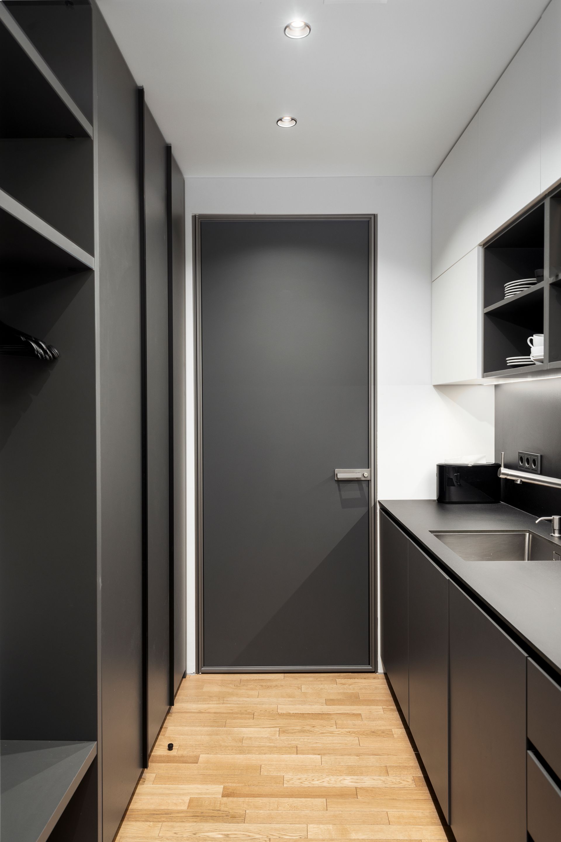 Ilgas koridorius, vedantis į virtuvę su juodomis spintelėmis ir juodomis durimis.