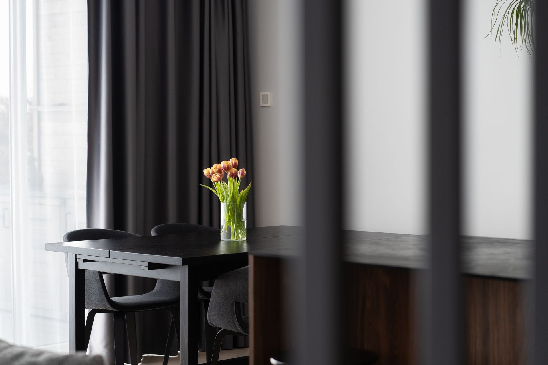 Kambarys su stalu, kėdėmis ir ant stalo stovinčia vaza su gėlėmis.