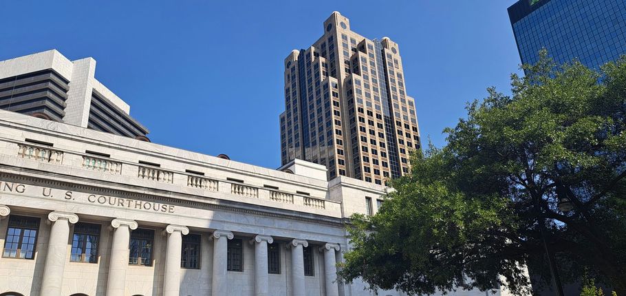Historic stone building and tall skyscraper under a clear blue sky, with green trees in the foreground.