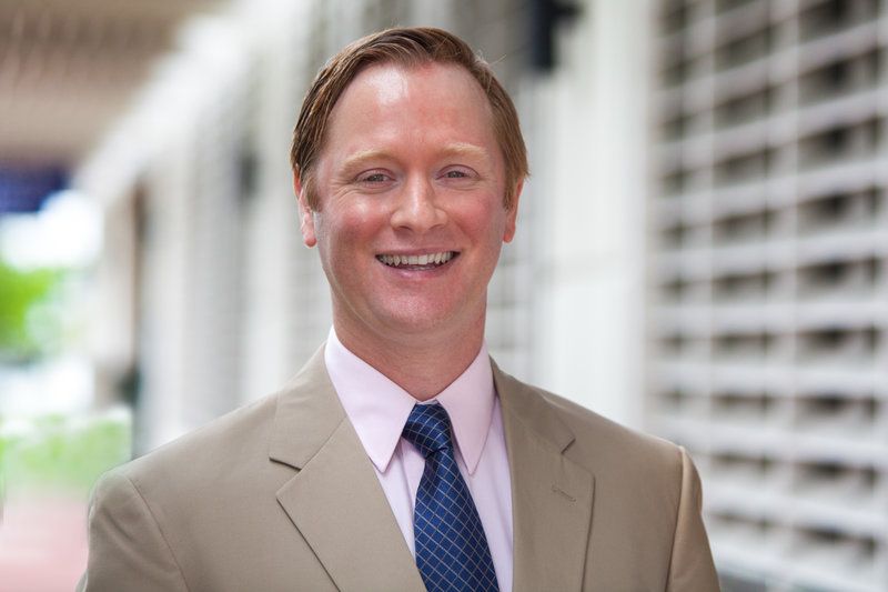 Smiling man in a tan suit and blue tie standing outdoors by a modern building