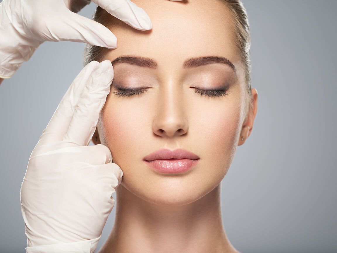 Gloved hands gently apply cosmetic treatment to a woman’s face with eyes closed