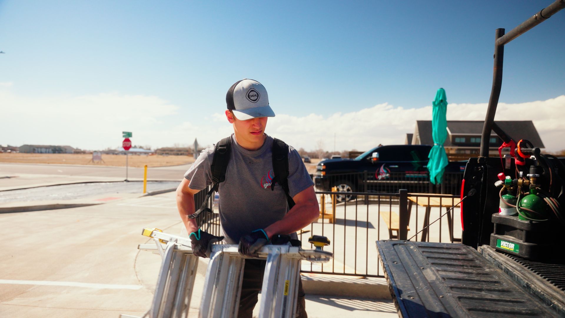 Person at an airport tarmac loading luggage beside a luggage cart under a clear blue sky