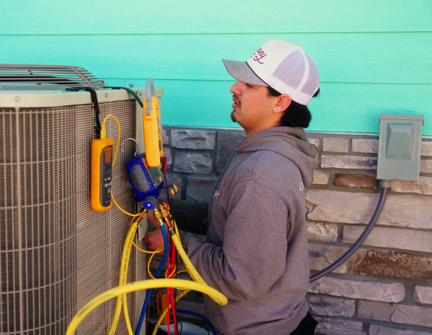 Technician servicing an outdoor HVAC unit with hoses and tools beside a brick wall