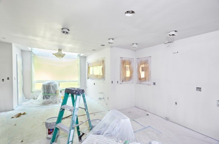 Bright white room under renovation with ladder, plastic-covered furniture, and taped windows