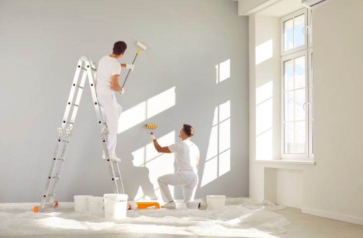 Two people painting a bright room white, one on a ladder and one kneeling near paint cans