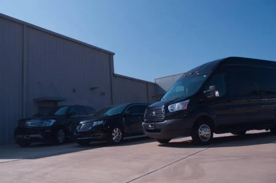 Black vehicles parked outside a light gray building under a clear, blue sky. A large van is in the foreground.