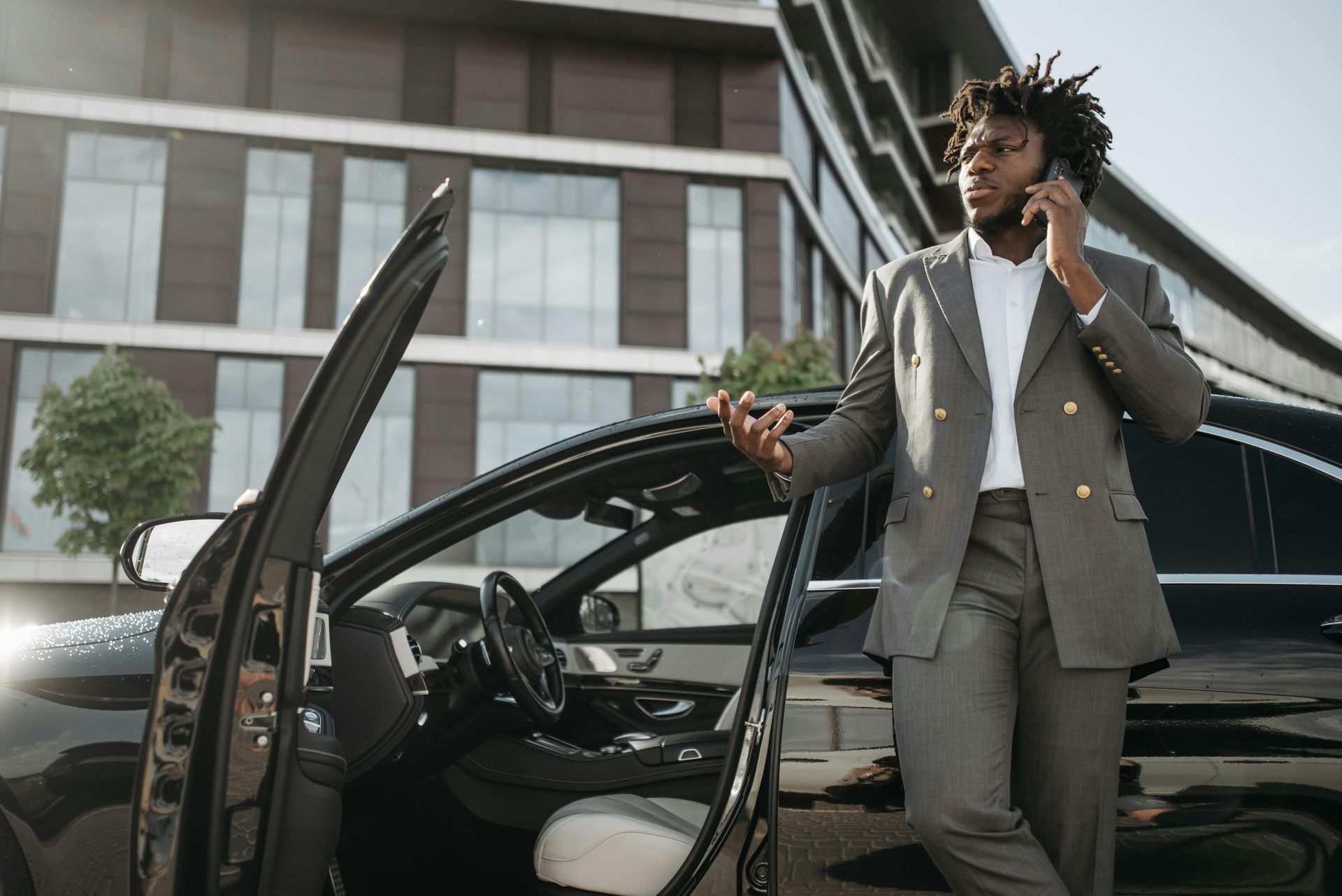 A Black man in a gray suit talks on the phone next to a black car with an open door in front of a modern building.