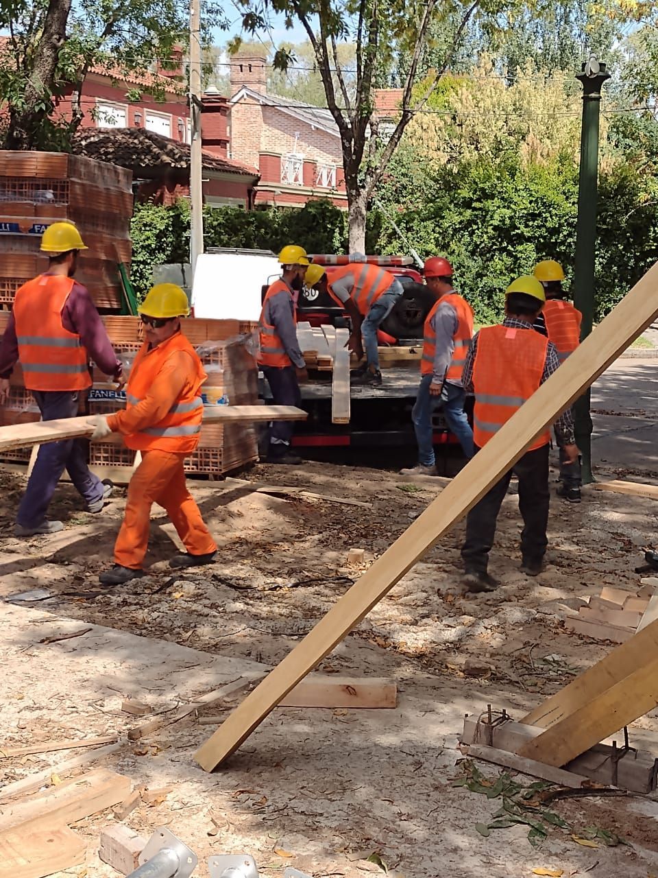 Obreros de la construcción con chalecos naranjas y cascos descargando materiales al aire libre.