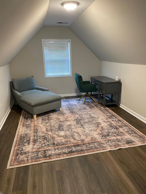Attic room with a chaise lounge, desk, and area rug. Neutral tones, sloped ceiling, and window.