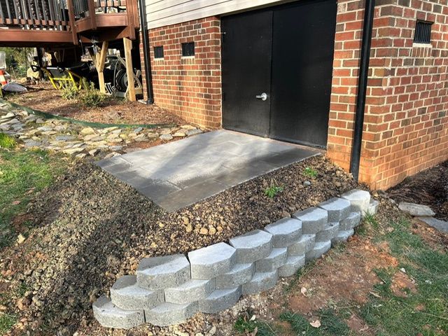 Brick building with black door. Stone retaining wall in front, dirt and grass surround.