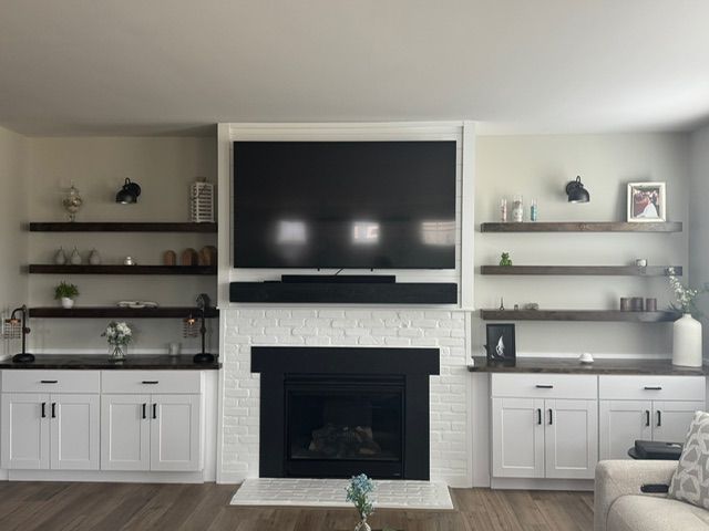 Living room with fireplace, white brick surround, black trim, large TV, white cabinets, and floating shelves.
