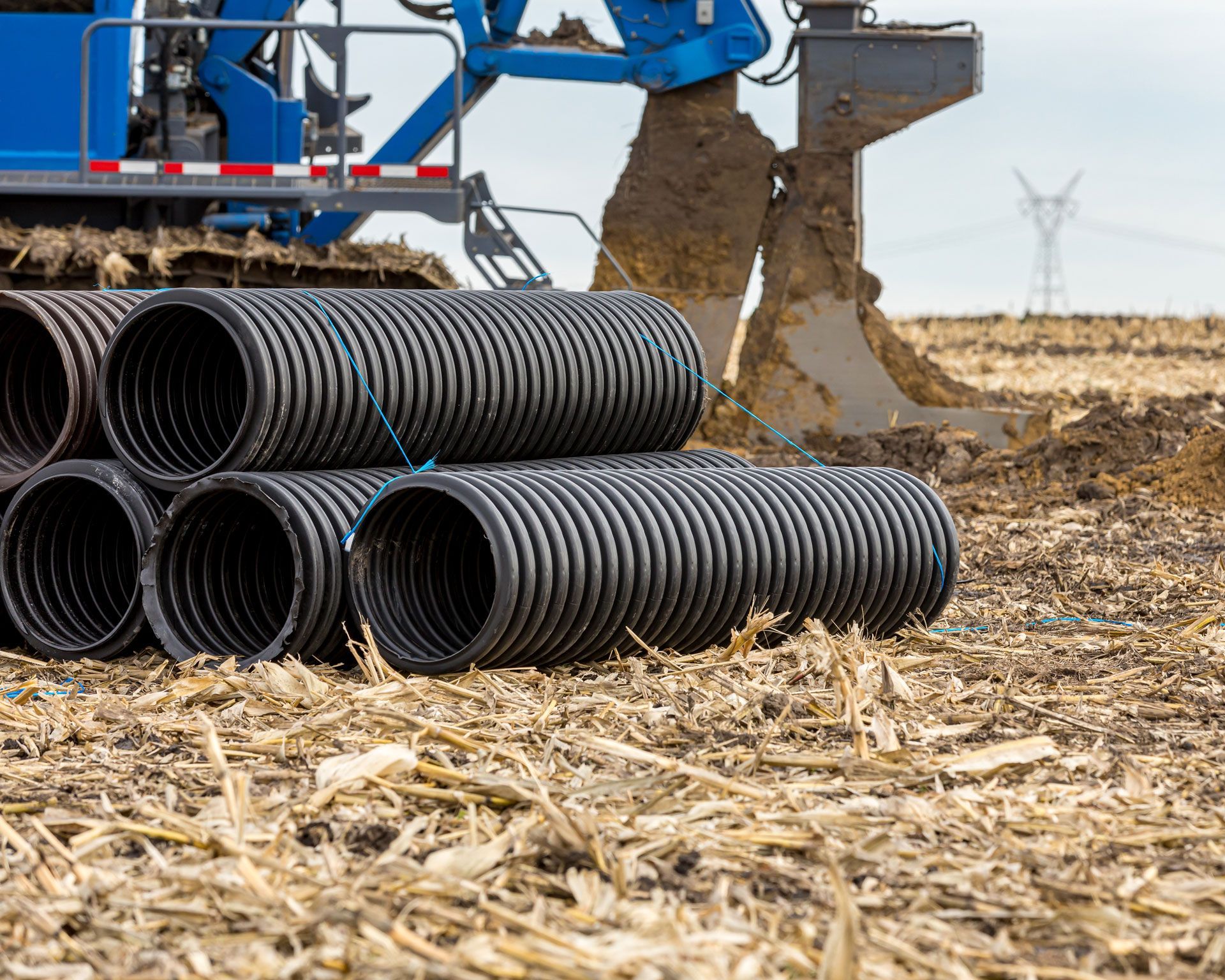 Black corrugated drainage pipes stacked near a blue trenching machine in a field.